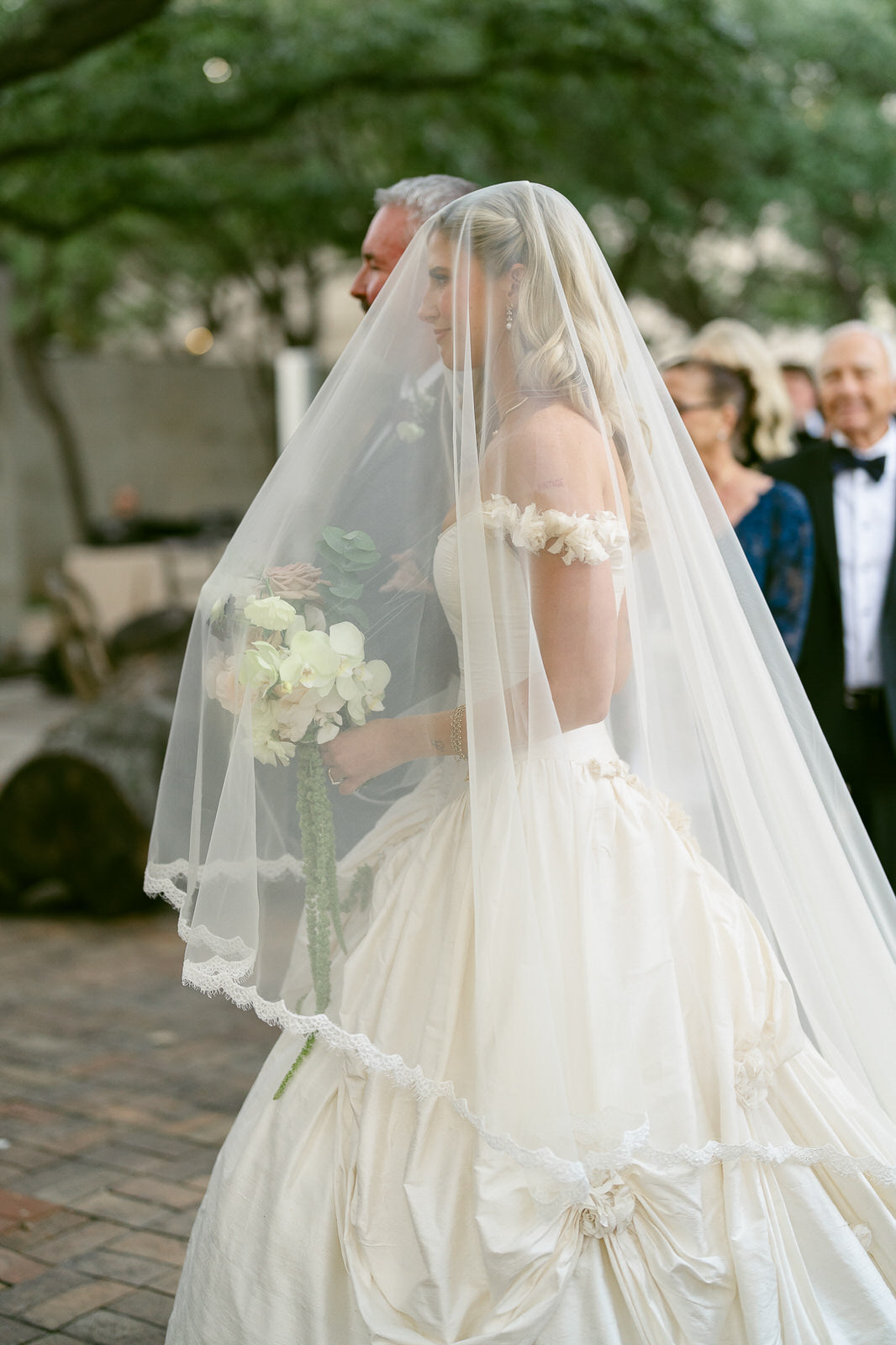 blonde bride in a vintage wedding dress with a long silk blusher veil, holding flowers, outdoors.