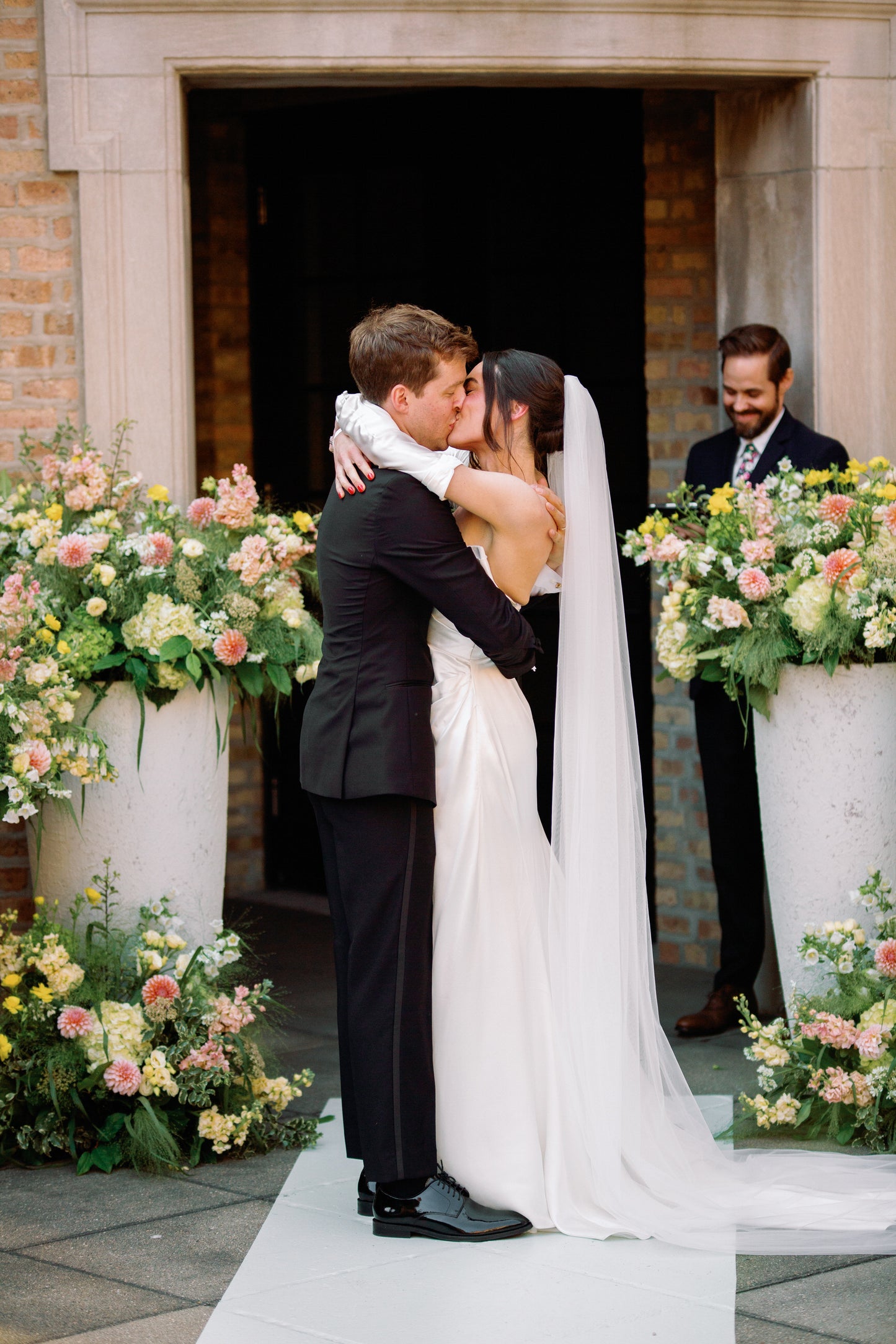 engaged Couple embracing in front of a building with pink and yellow loral arrangements with bride in fingerless crepe bridal glove sleeves