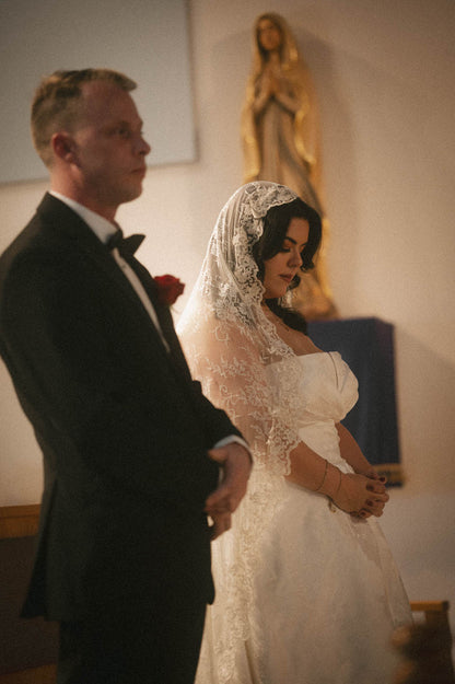 Bride and groom standing in an orthodox church with bride in strapless gown and mantilla bridal veil in lace with religious statue in the background.