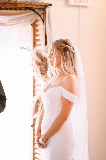 bride in off shoulder ruched wedding dress with a simple fingertip bridal veil standing against pampa grass alter 