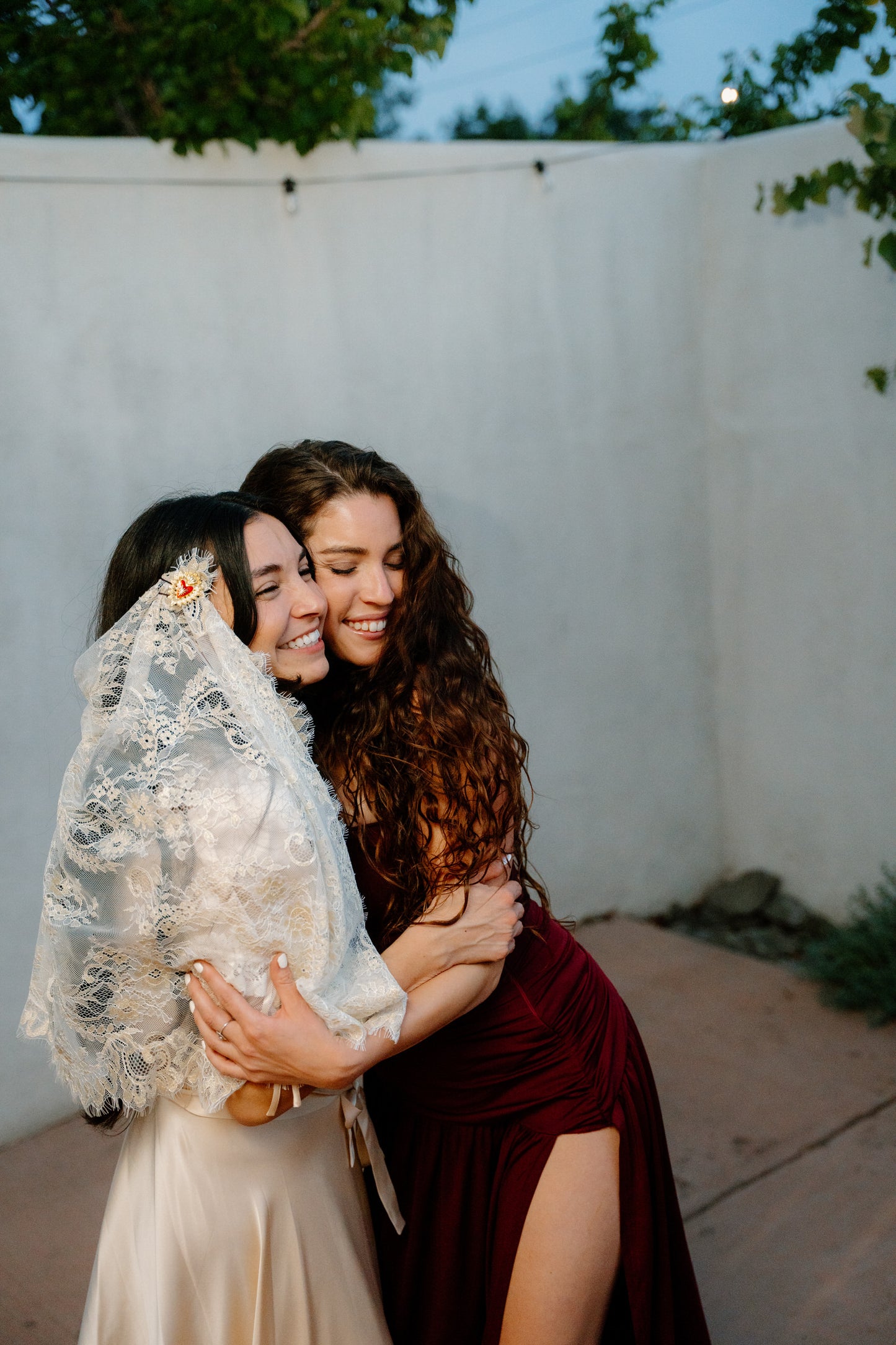 bride and maid of honor hugging, one in a champagne dress with draped lace bridal veil elbow length