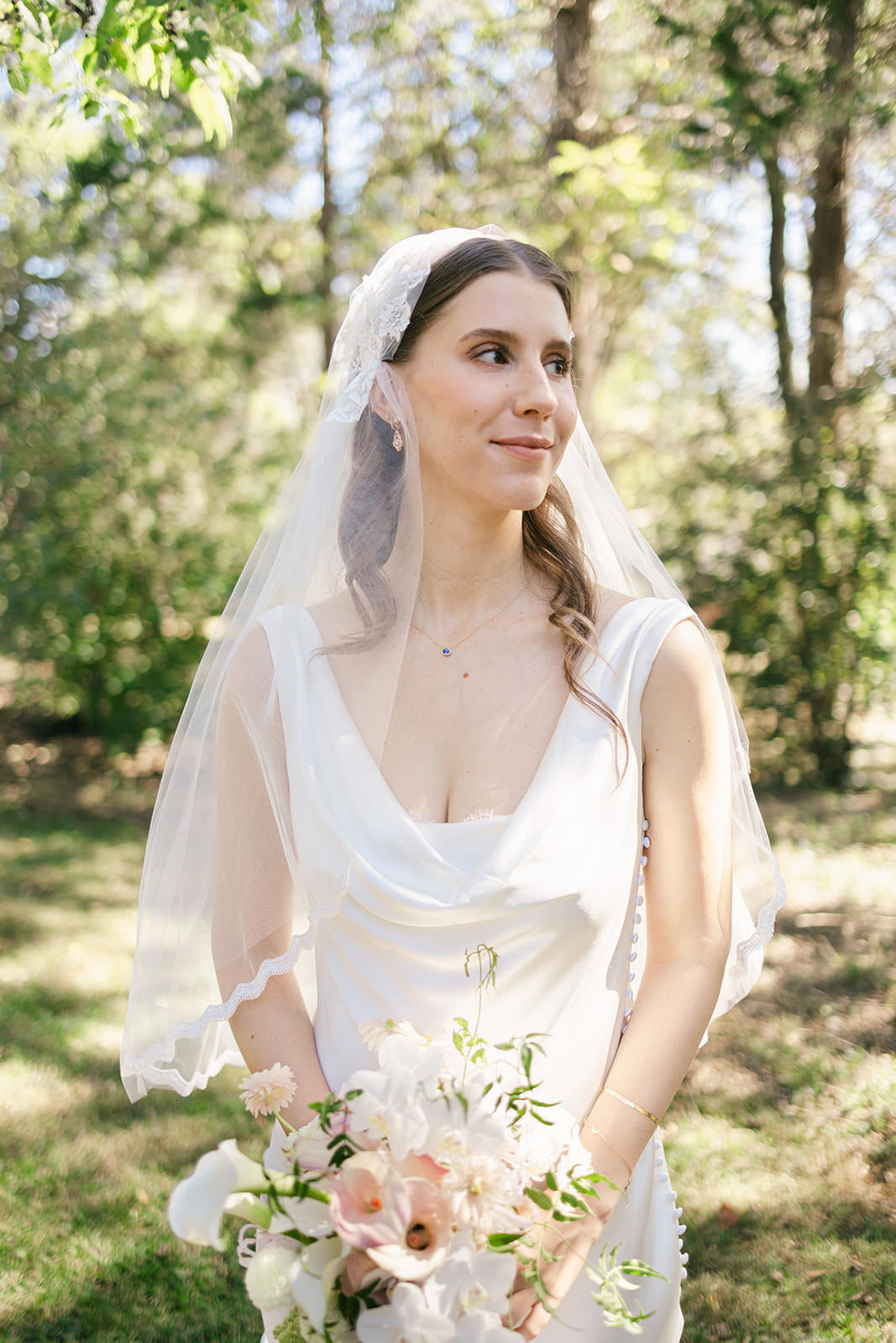 Brunette bride in a white wedding dress with a Juliet cap wedding veil holding a bouquet outdoors.