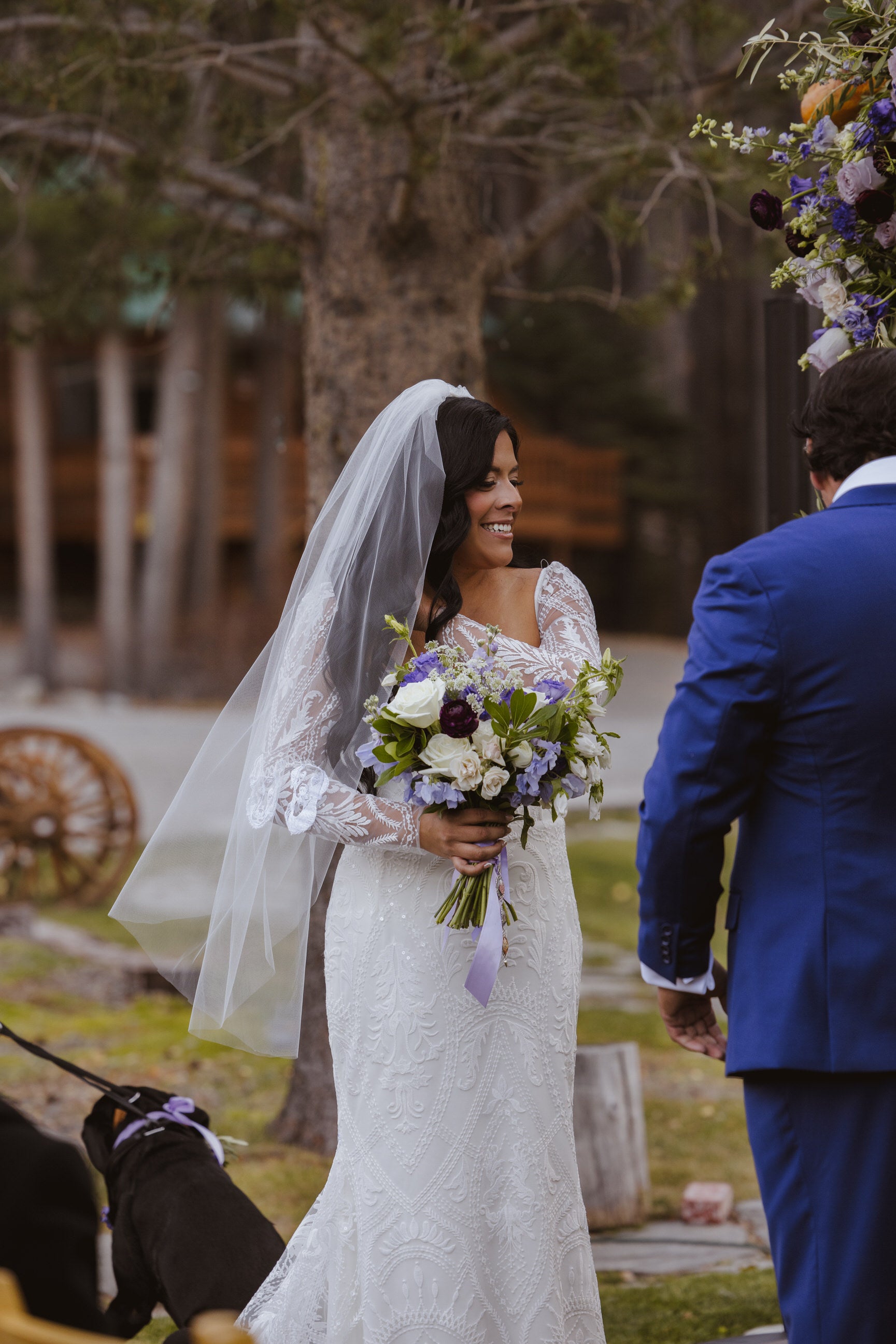 Bride in a beaded long sleeve wedding dress holding a bouquet, standing outdoors with a fingertip veil with embroidered dog design