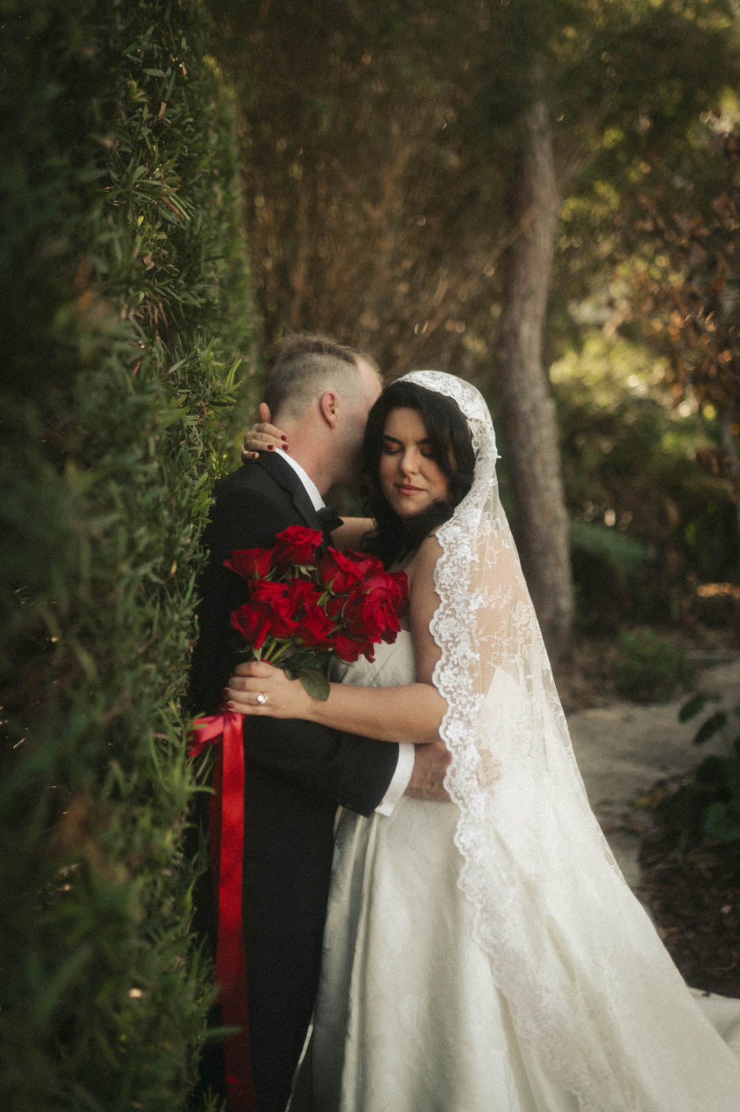 Wedding couple embracing in a forest setting with a bride  in lace mantilla knee length veil holding red rose bouquet.