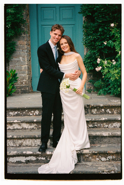 engaged couple standing on stone steps with bride in cat eye neck gown and scarf in front of a teal door with greenery.