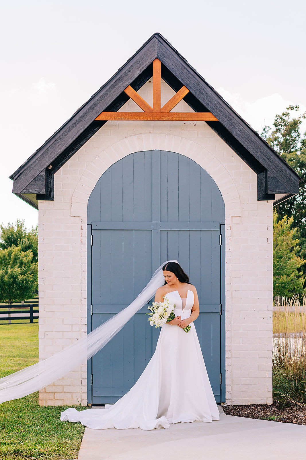 bride in a white notch neckline dress and wedding veil in simple tulle standing in front of a small building with a blue door.