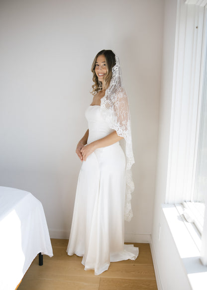 bride in a white strapless wedding dress with a sheer chantilly lace mantilla veil over low curled hair standing in a room with a bed and window.