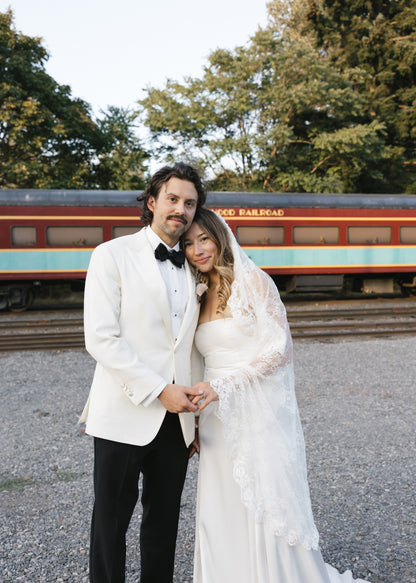 Bride and groom in wedding attire standing in front of a vintage train. bride wearing ballet length chantilly lace veil from one blushing bride