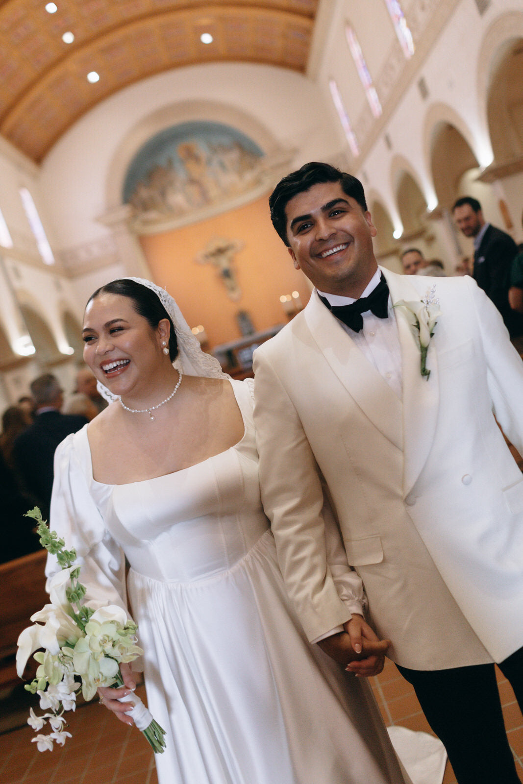 A smiling bride in a white satin shoulder wedding gown holding a bouquet of white lilies walks hand-in-hand with a groom in a cream tuxedo and black bow tie inside a church.