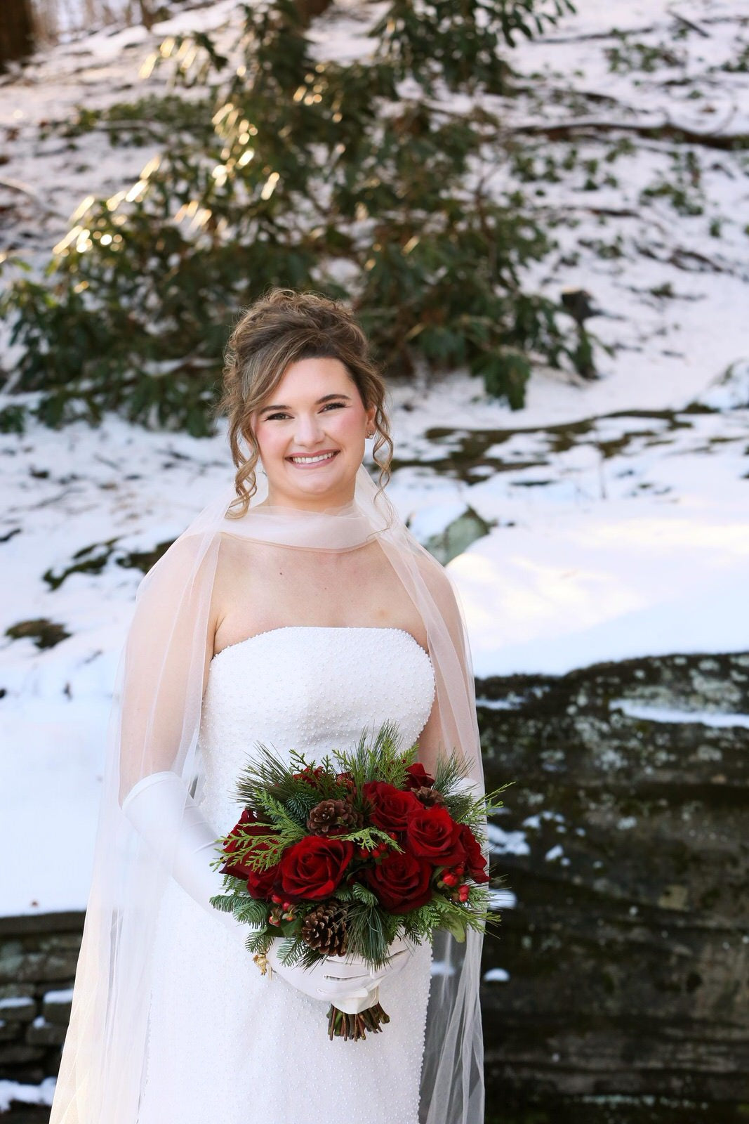 Bride wearing veil under high updo and tulle scarf and satin gloves  holding a bouquet of red flowers in a snowy outdoor setting