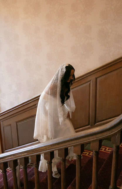 Bride in a white off shoulder bridal dress and lace fingertip veil walking up historical staircase in UK