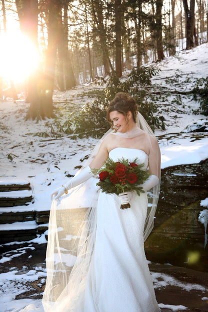 Bride in a snowy forest holding a bouquet of red flowers wearing satin gloves, bridal neck scarf and matching long chapel veil