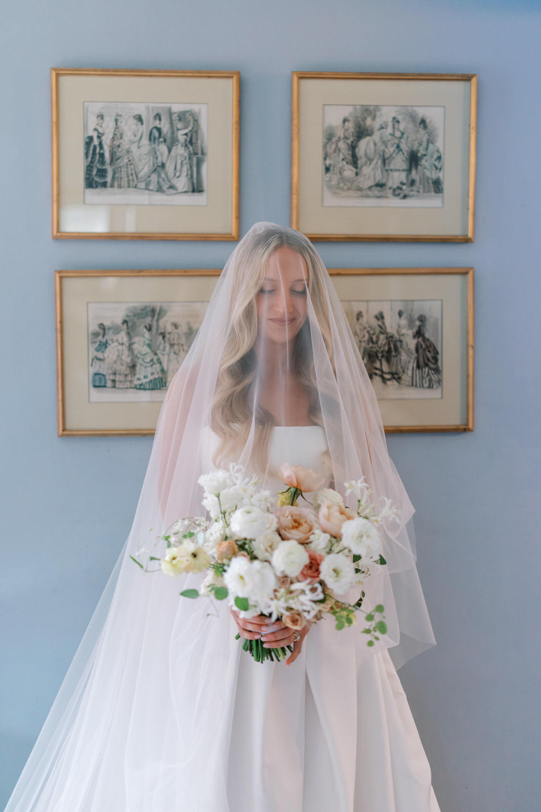 Bride wearing a drop blusher wedding veil holding a bouquet in front of a wall with framed artwork