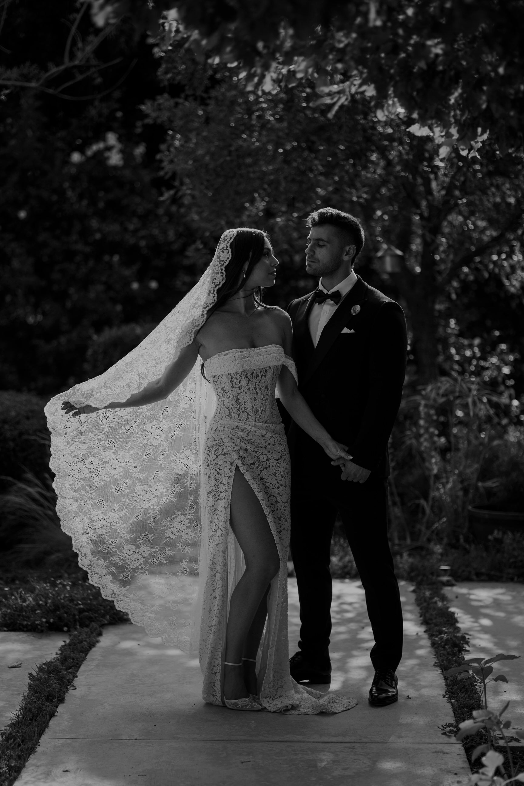 elegant floor length mantilla lace veil on bride in strapless lace gown with slit admiring her groom