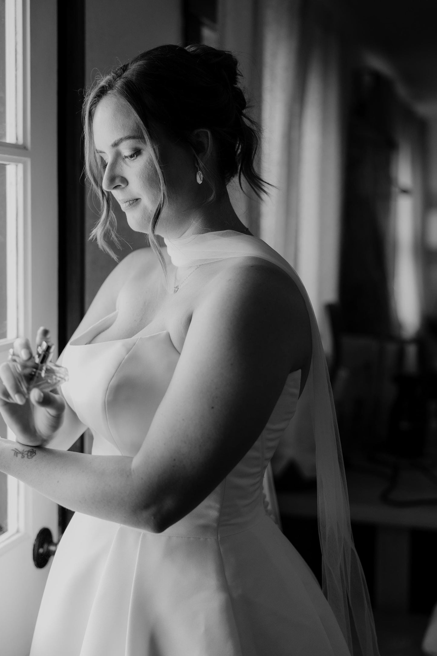 Black and white photo of a bride in a wedding dress and narrow tulle bridal scarf standing by a window.