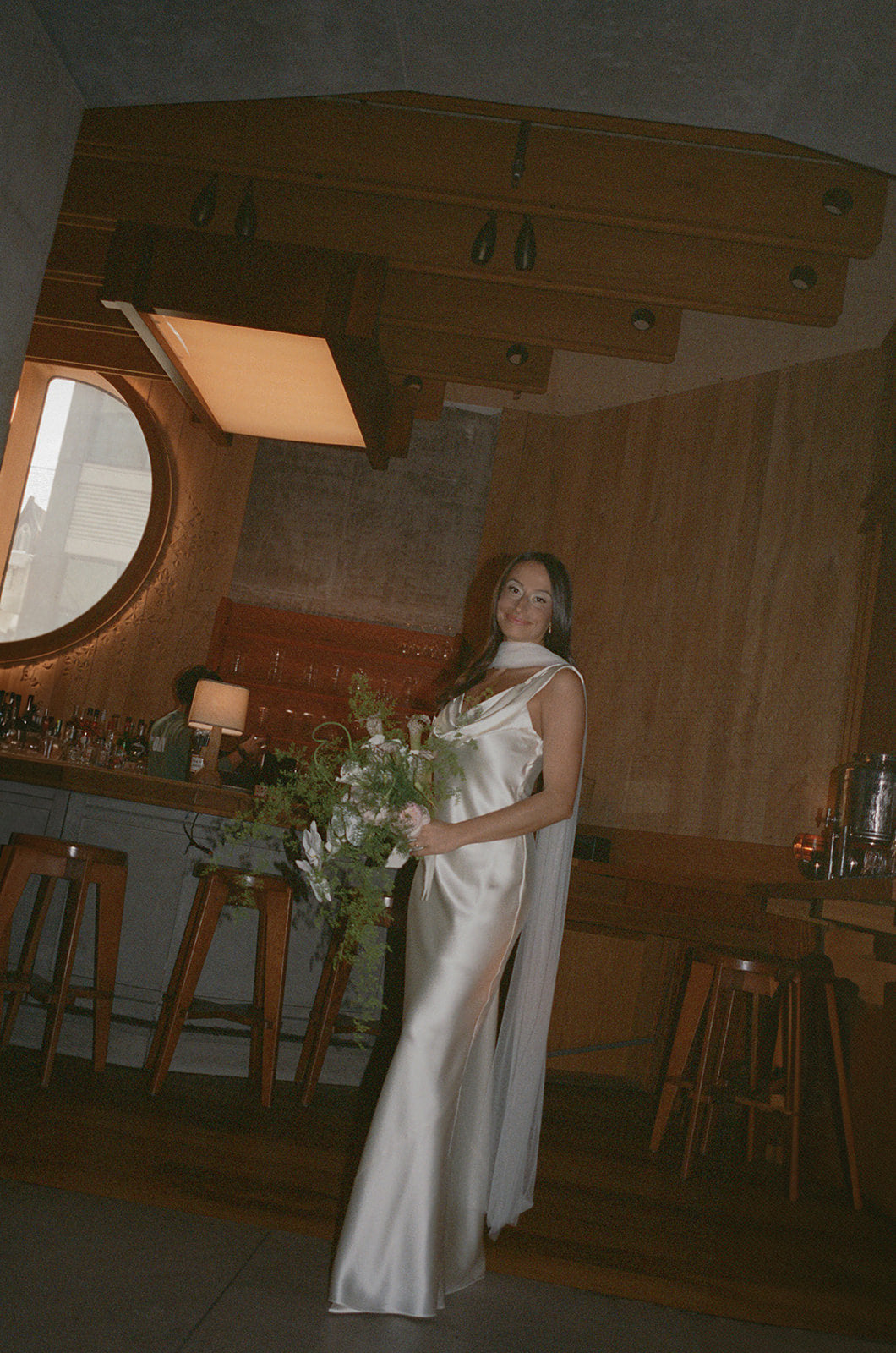 Simple glam bride in a white bridal dress holding flowers in a rustic interior setting wearing a neck tulle scarf