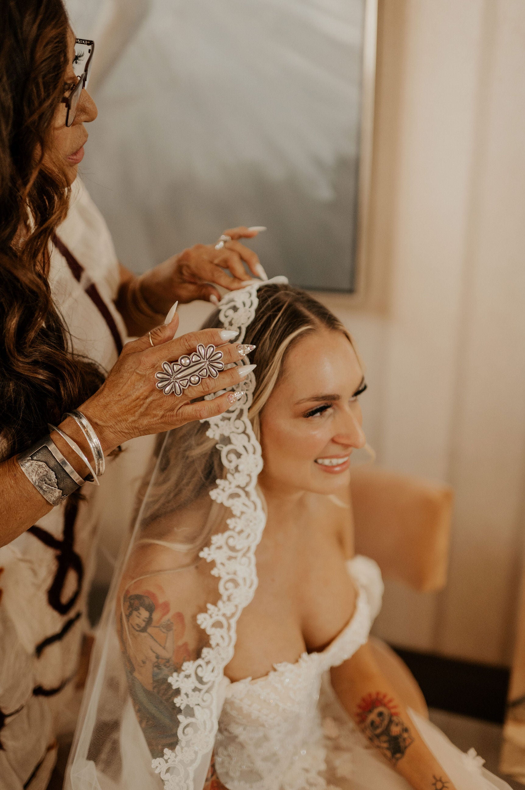 stylist positioning the mantilla wedding veil on top of curled hair down as bride smiles